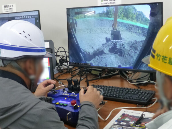 無人化施工機械操作体験会。協力業者の操作体験を竹花組社員が操作の仕方を教えている様子の画像。
