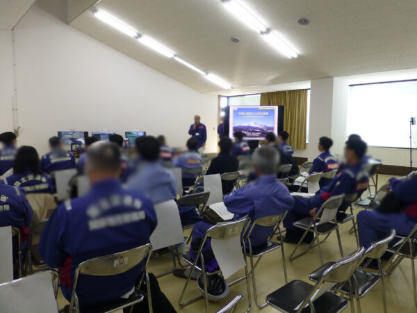 無人化施工機械操作体験会の1日目。浅間出張所内の職員さん向けの体験会。青い作業服を着た人がずらりと椅子に座っているのを最後列から部屋全体を撮影した画像。最前列の前には司会進行役の方が立って話している様子。