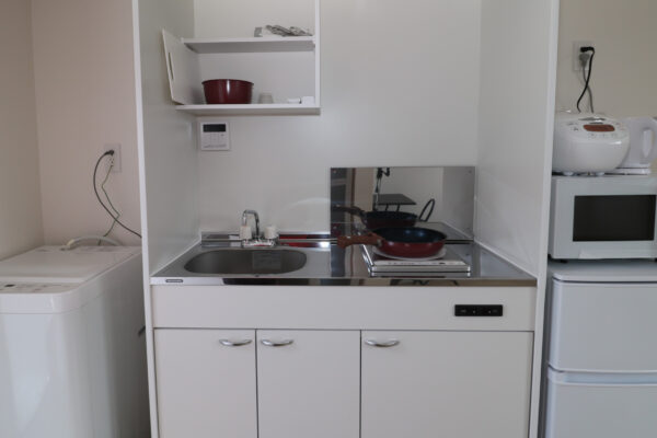 住宅の内観。洗濯機、フライパンが乗ったキッチン、冷蔵庫、レンジ、湯沸かしポット、炊飯器が写っている画像